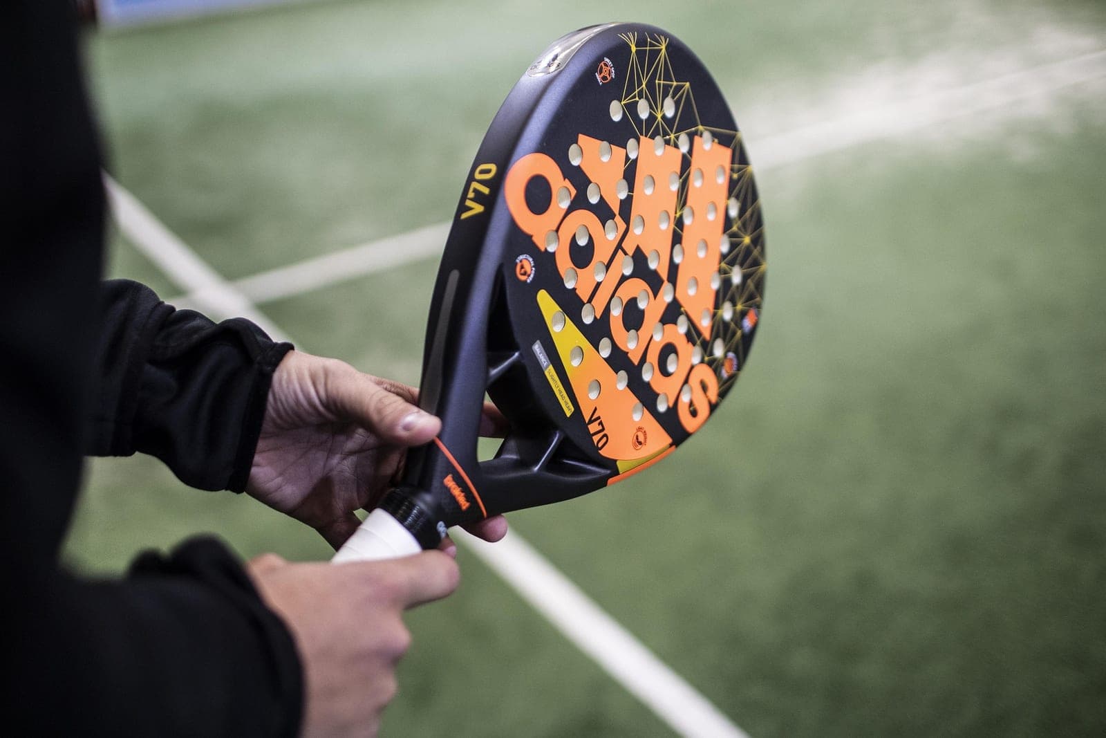 Hand holding an orange Adidas V70 padel racket from below, indoor astroturf court in the background