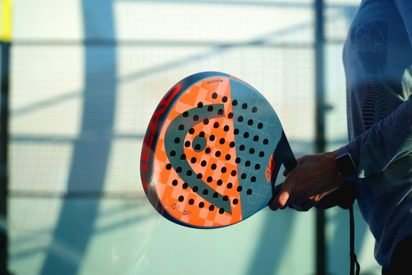 Orange and grey HEAD Evo padel racket held against an outdoor padel court's glass wall in low sunlight