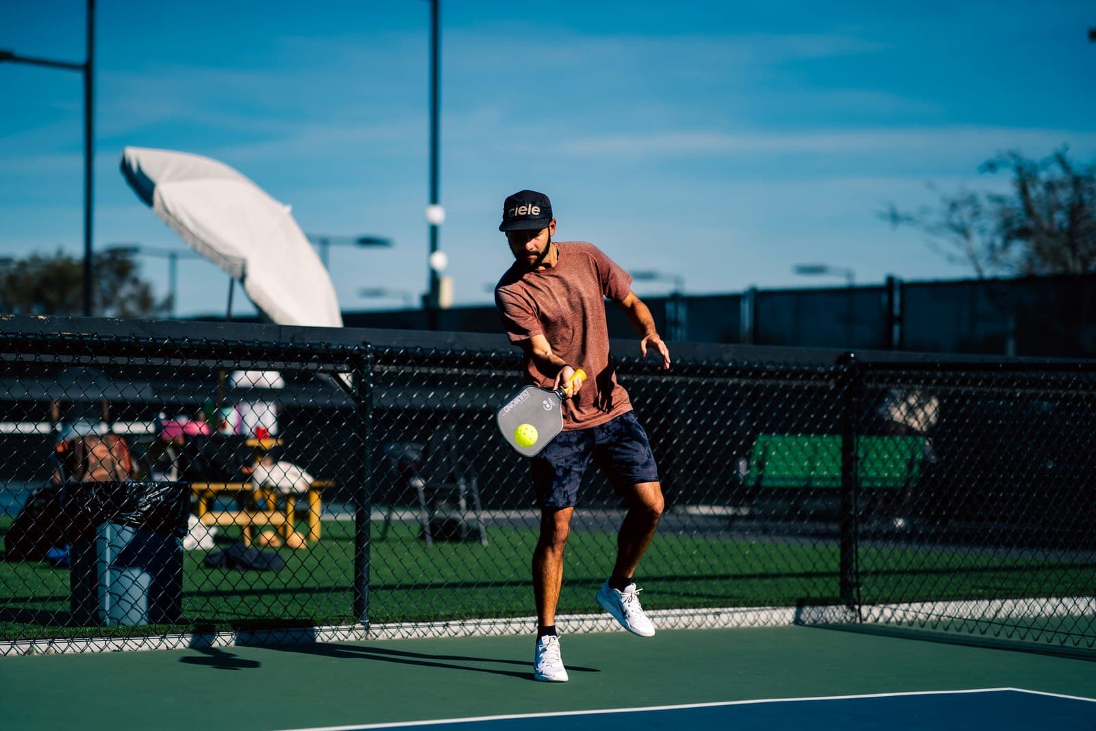 Male pickleball player mid-swing striking a yellow ball on an outdoor court under a sun shade