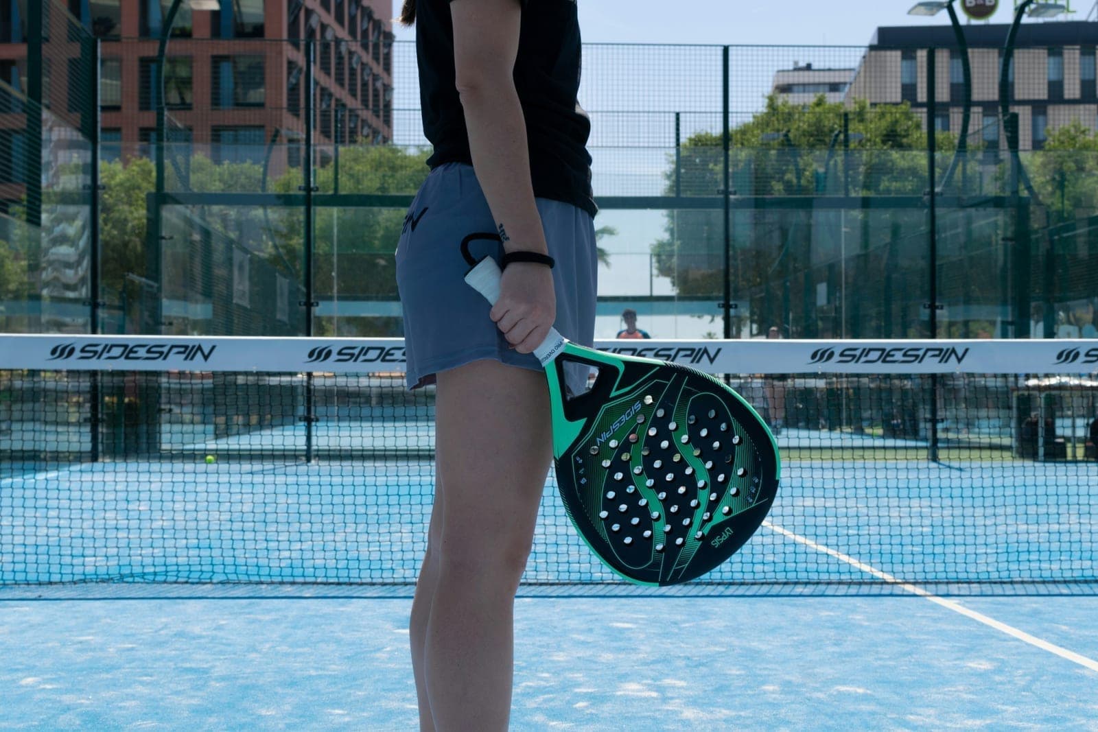 Female padel player from behind on a blue outdoor court holding a green Sidespin Lyysis racket