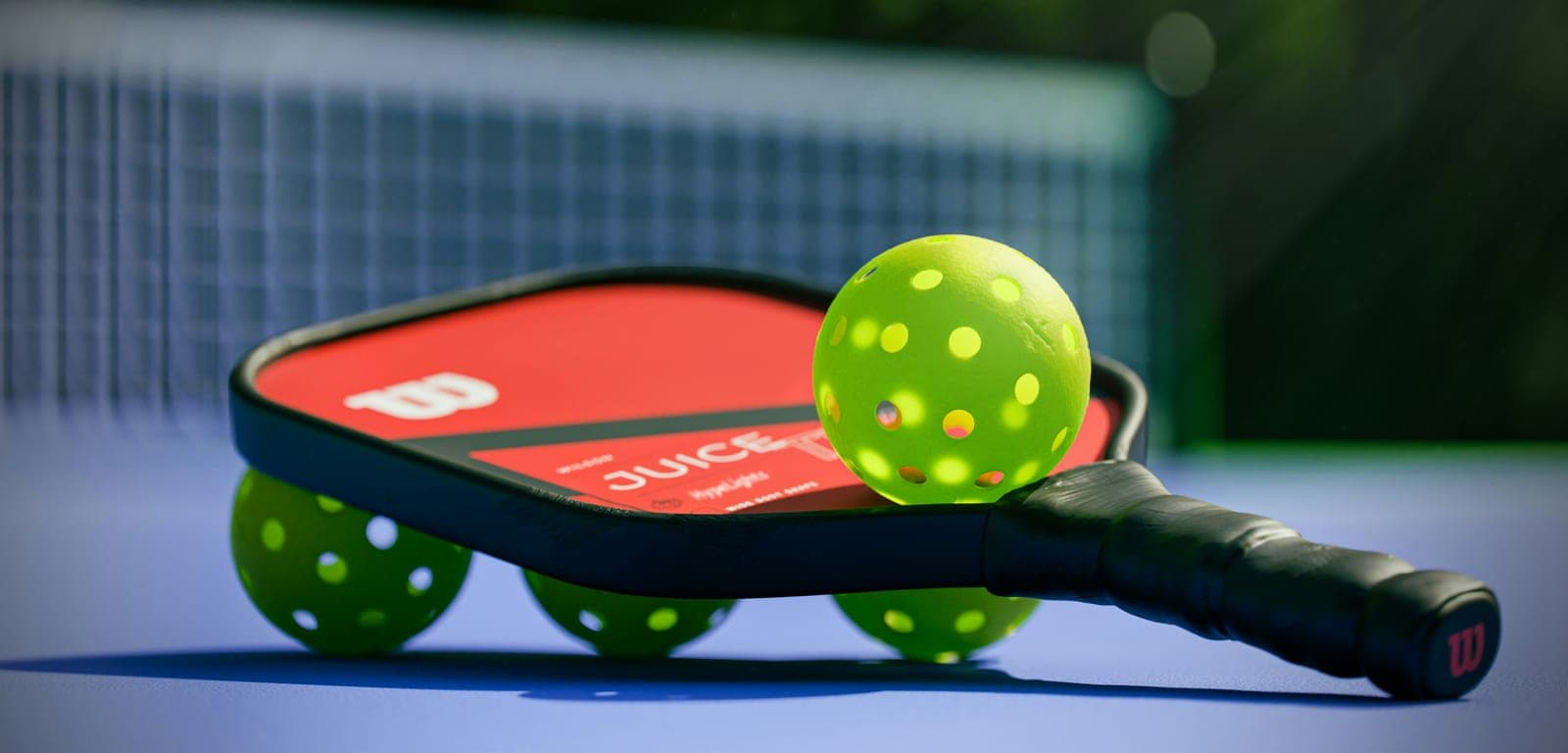 Wilson Juice pickleball paddle with three lime green pickleballs on a blue court, net behind