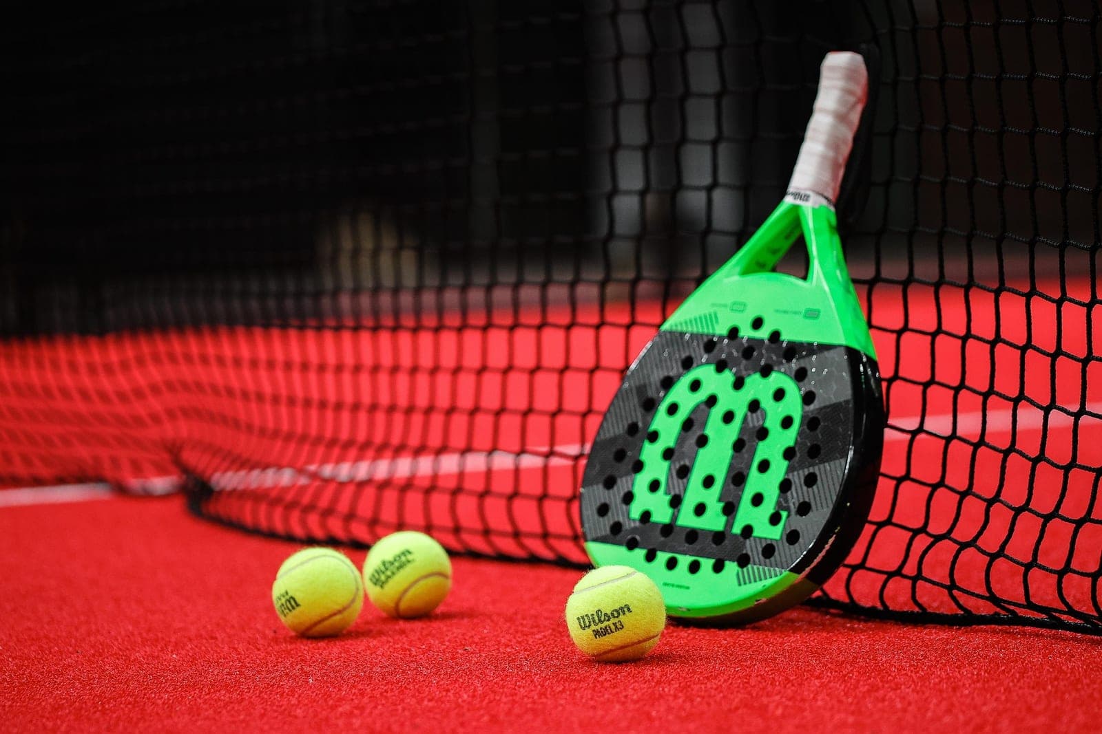 Green and black Wilson padel racket against a net on a red court with three Wilson Padel X3 balls in front