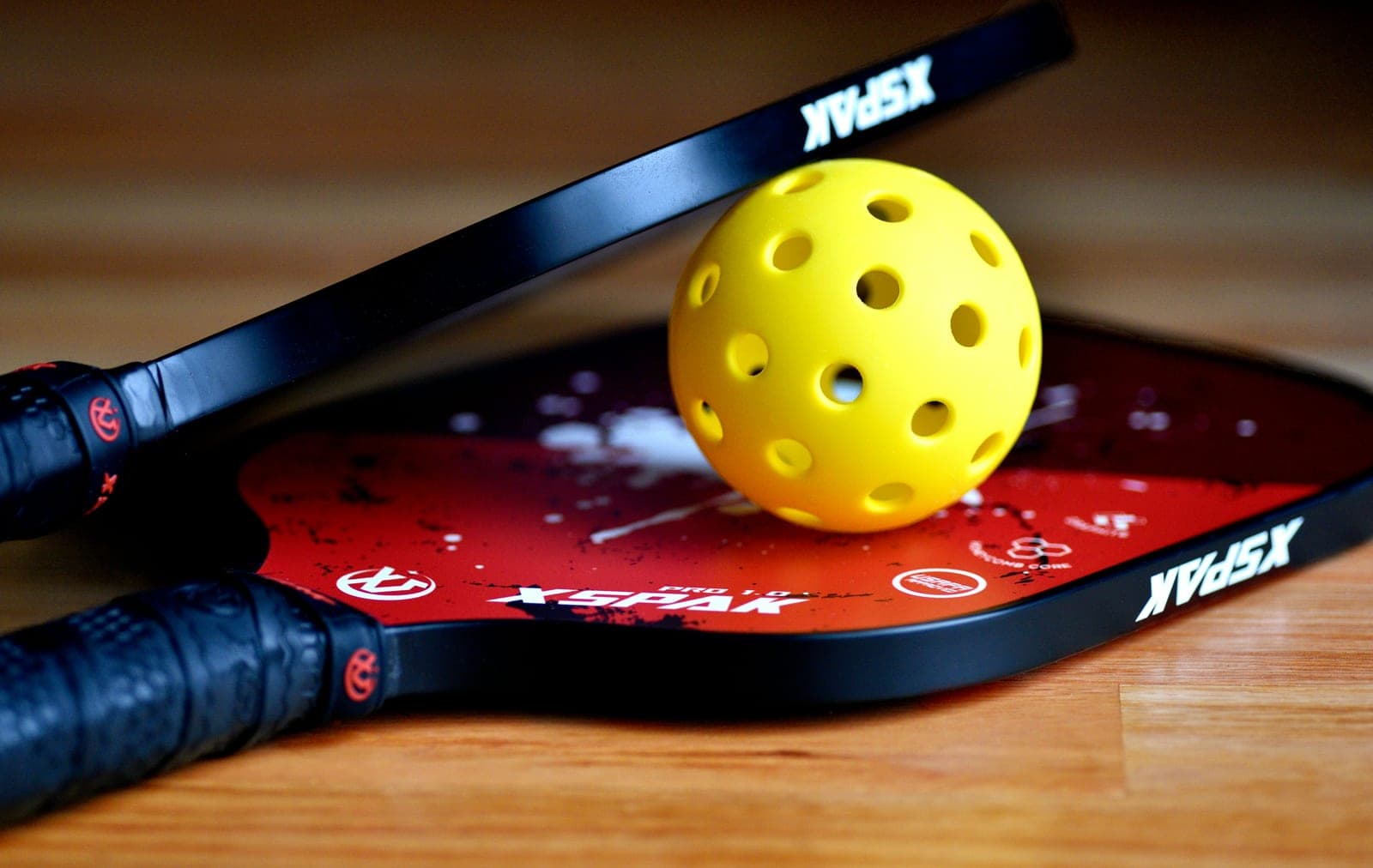 Two black and red XSPAK pickleball paddles crossed with a yellow pickleball on a wooden floor